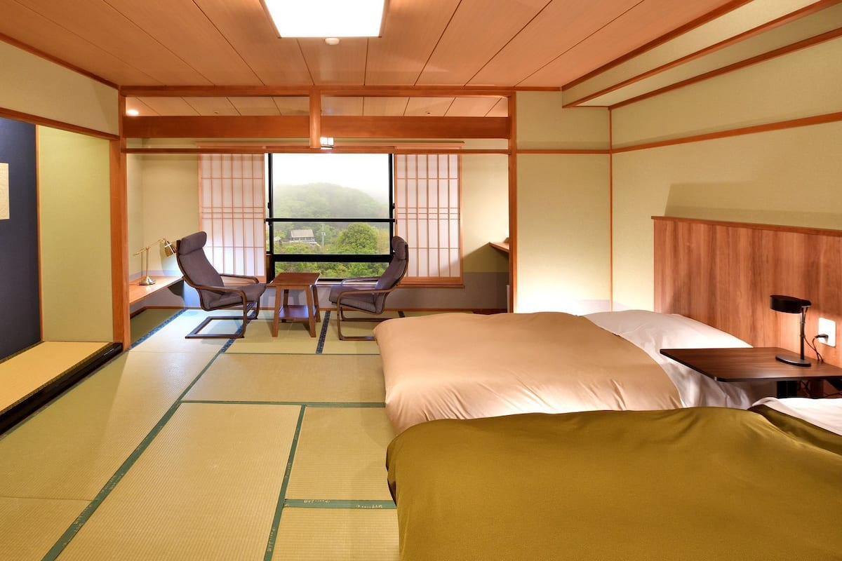 A traditional Japanese-style hotel room with tatami flooring, a bed, a desk, and a view of the outdoors.