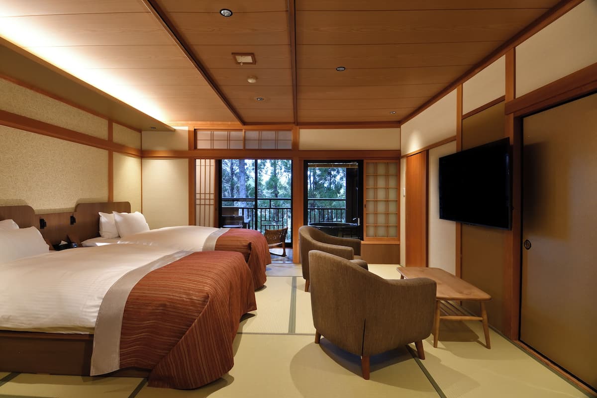 A hotel room with a large bed, two armchairs, a small table, a TV, and a balcony with a view of trees.