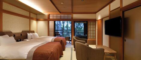 A hotel room with a large bed, two armchairs, a small table, a TV, and a balcony with a view of trees.