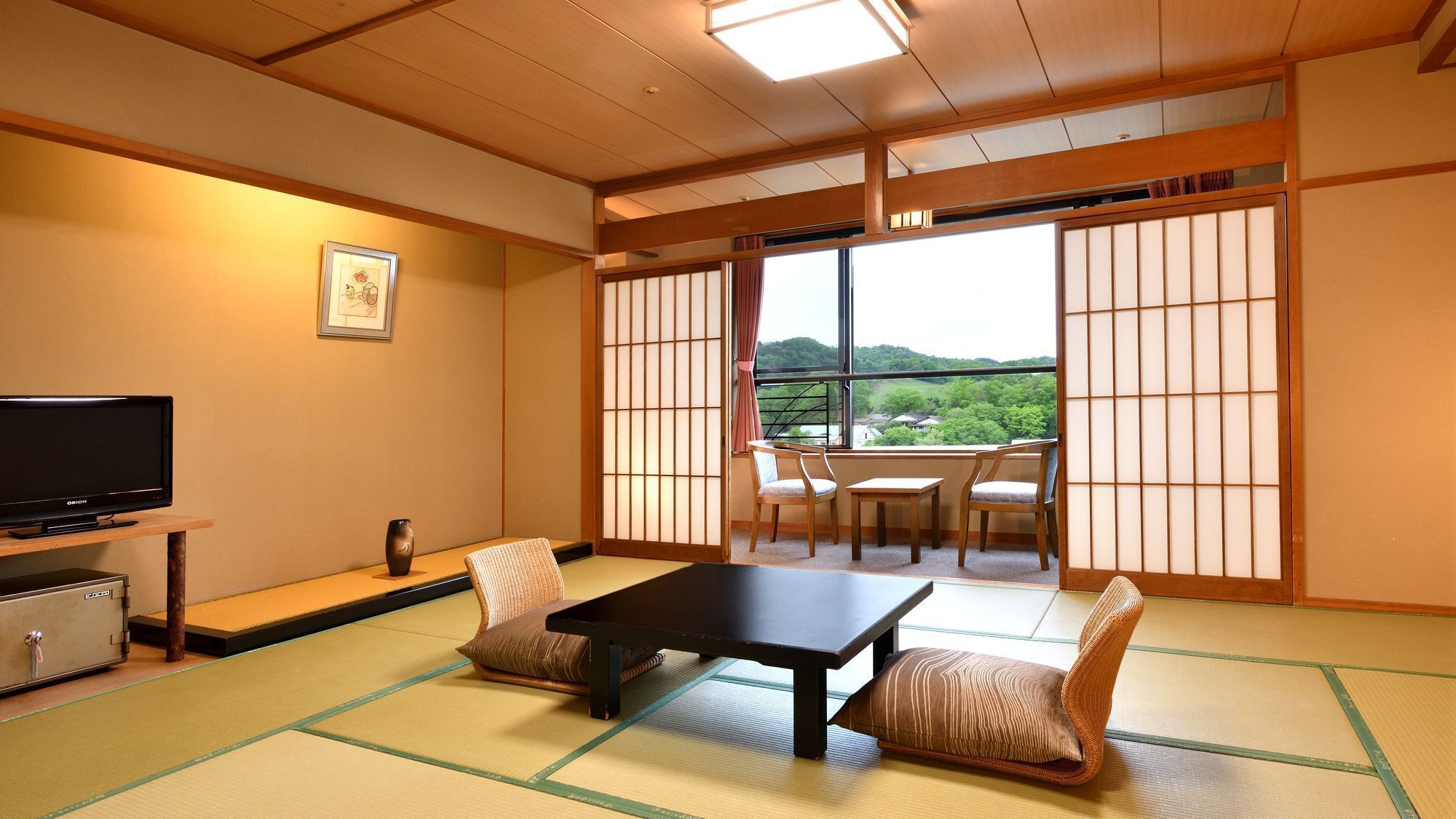 A traditional Japanese room with tatami flooring, a television, a table, chairs, and a view of a green landscape through shoji screens.