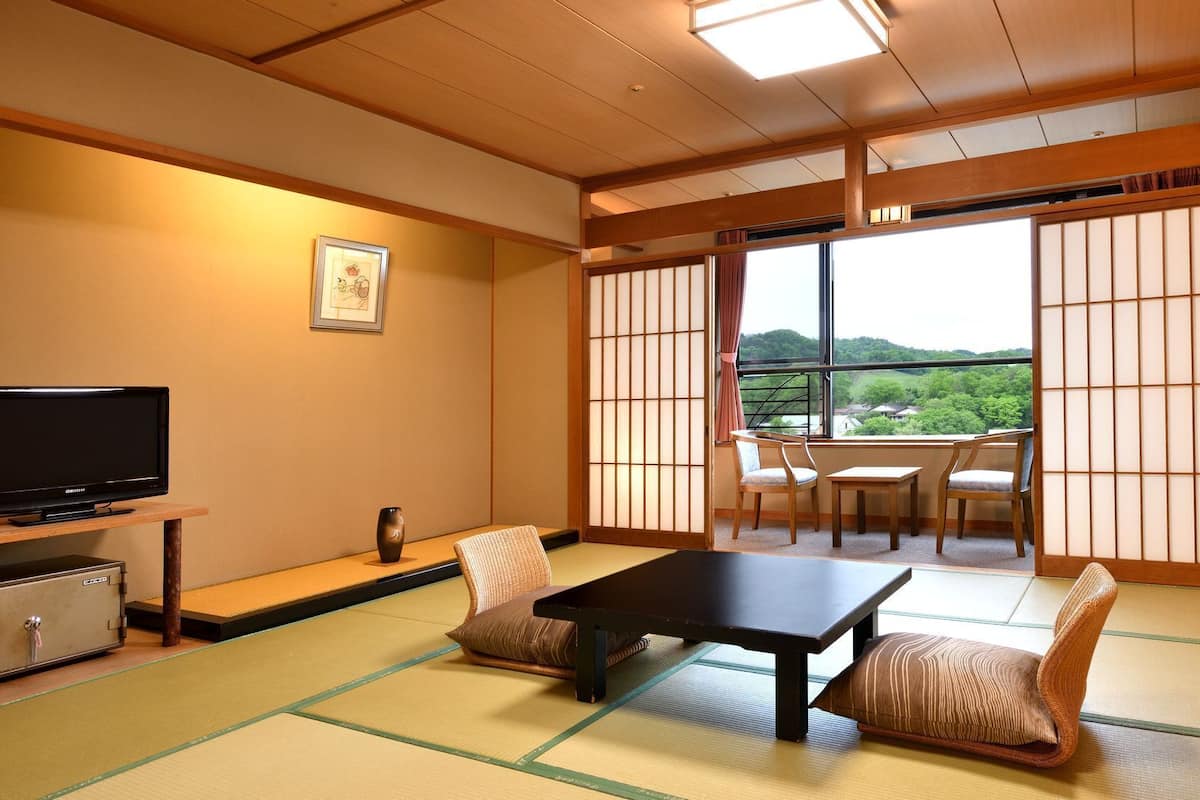 A traditional Japanese room with tatami flooring, a television, a table, chairs, and a view of a green landscape through shoji screens.