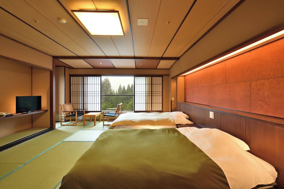 A hotel room with two beds, a television, a chair, and a view of trees through large windows.