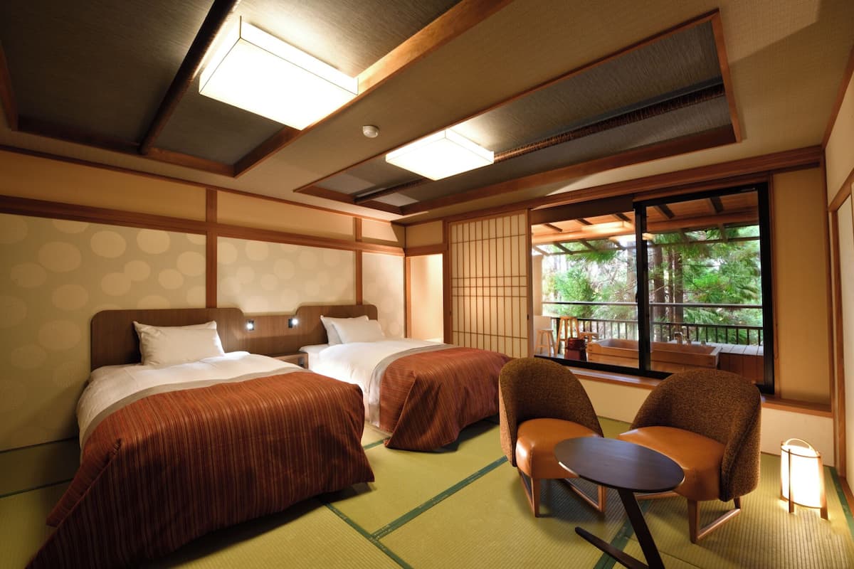 A hotel room with two beds, a small table, chairs, and a view of the outdoors.