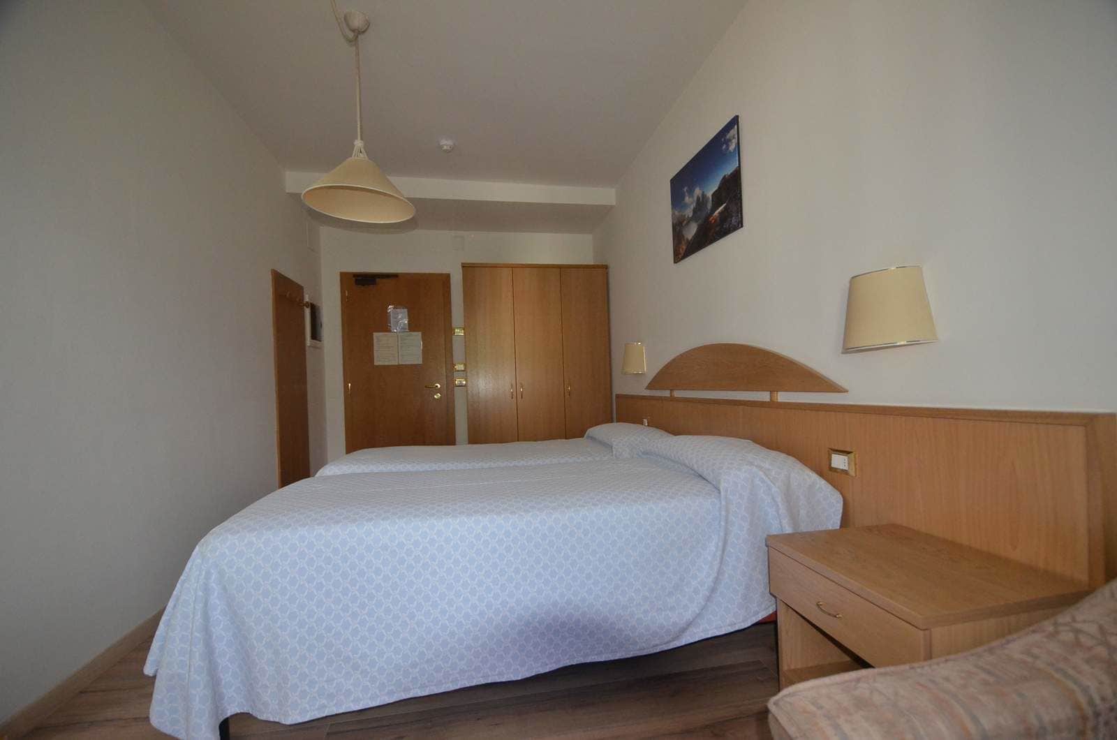 Ein Hotelzimmer mit Doppelbett, Nachttisch aus Holz, einer Nachttischlampe, einem Holzschrank und einer Tür mit einem Hinweis.