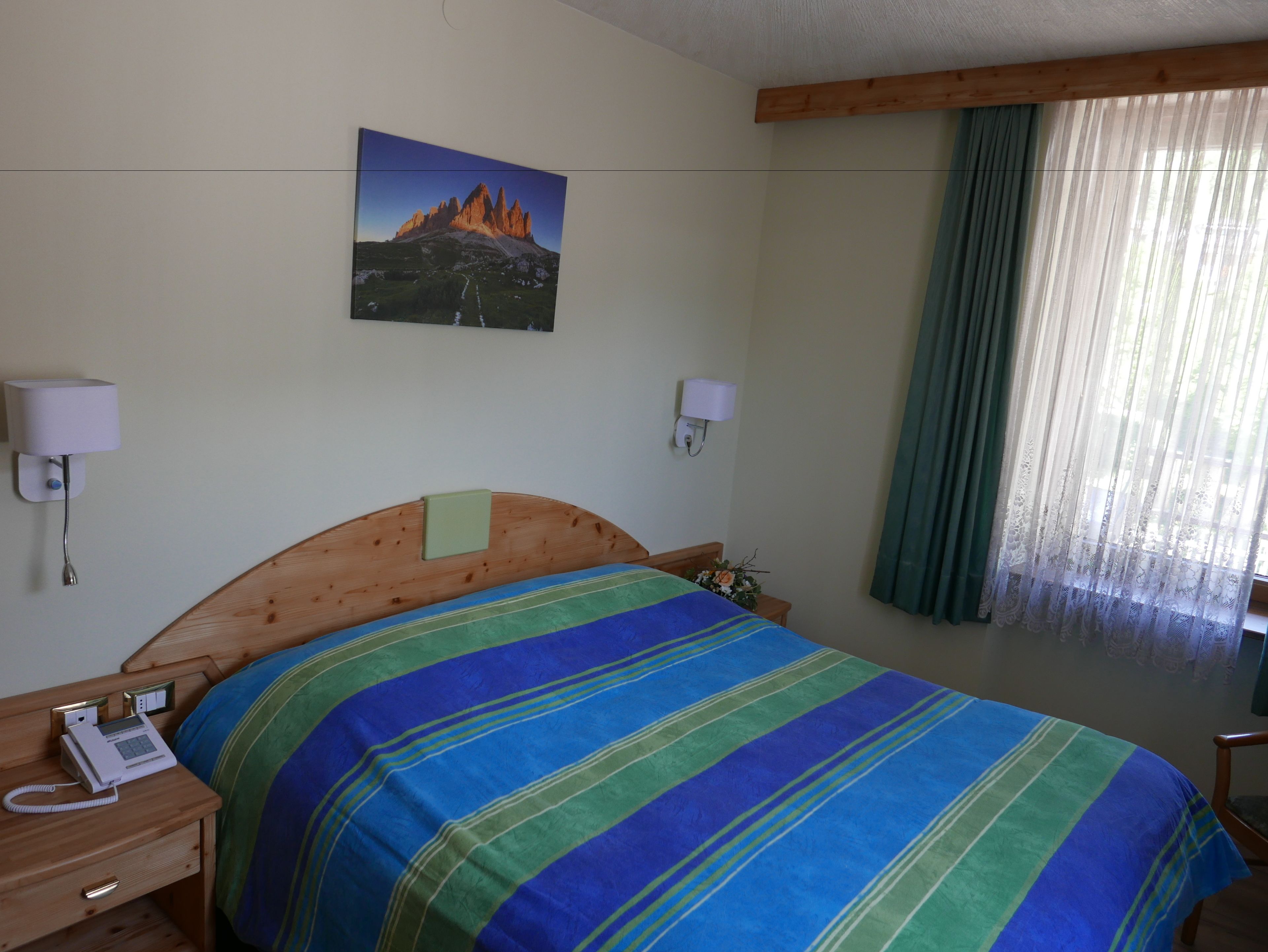 Ein Hotelzimmer mit einem hölzernen Kopfteil, einer gestreiften Bettdecke, einem Gemälde von einem Berg, einer Nachttischlampe und einem Telefon auf dem Nachttisch.