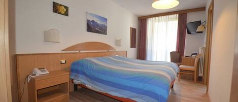 Habitación de hotel con cama, mesita de noche, lámpara, silla y ventana con cortinas.