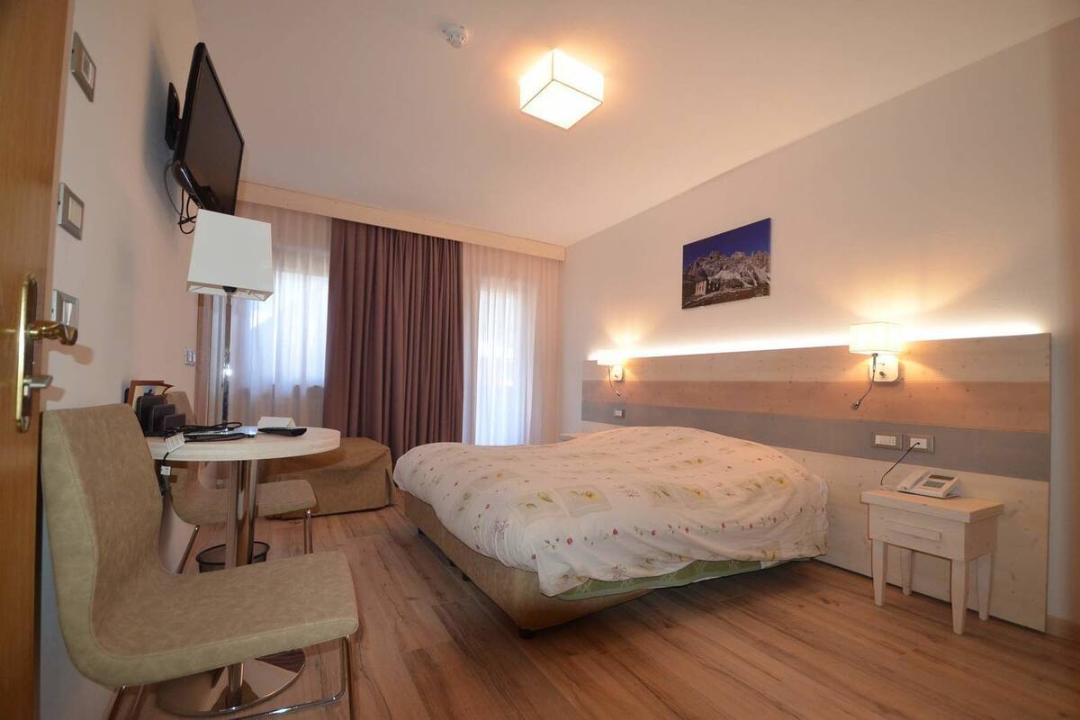 A hotel room with a bed, a desk, a chair, a TV, and a bedside table with a lamp.