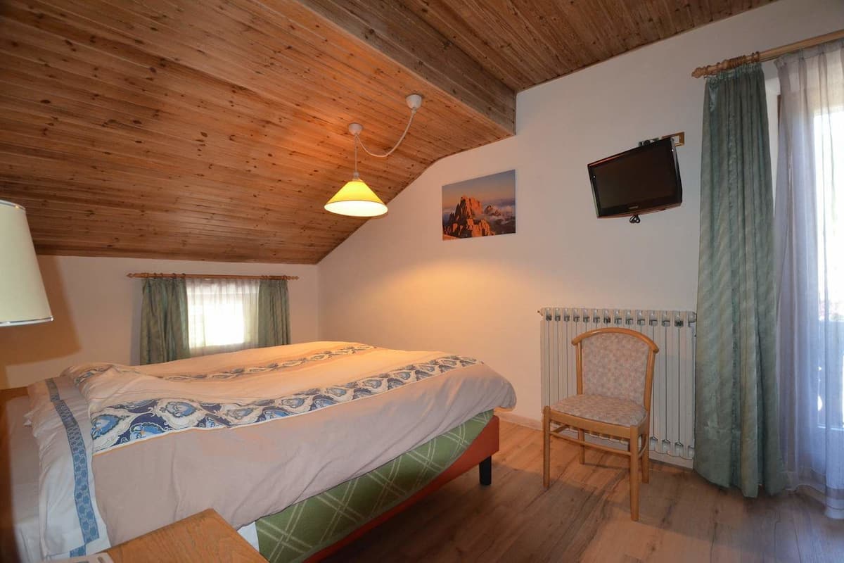 A bedroom with a wooden ceiling, a bed with patterned bedding, a chair, a TV mounted on the wall, and a window with curtains.
