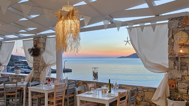 Un espace de restauration en plein air avec des murs en pierre, des rideaux blancs et une vue sur la mer au coucher du soleil.