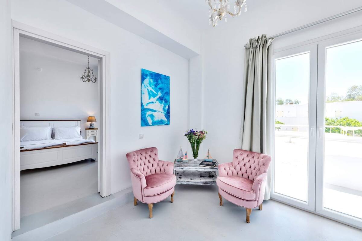 Una habitació lluminosa amb un llit, dues butaques roses, un quadre abstracte blau i una vista per una finestra gran.