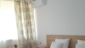 A hotel room with a large bed, a wooden headboard, white pillows, a nightstand, a small table with a vase of flowers, and a window with sheer curtains. - Semiramis Hotel (Saturn)