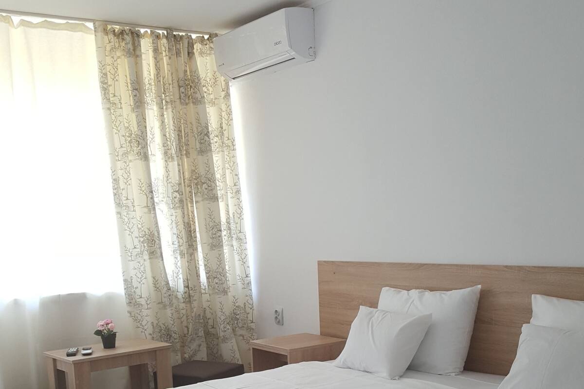 A hotel room with a large bed, a wooden headboard, white pillows, a nightstand, a small table with a vase of flowers, and a window with sheer curtains.