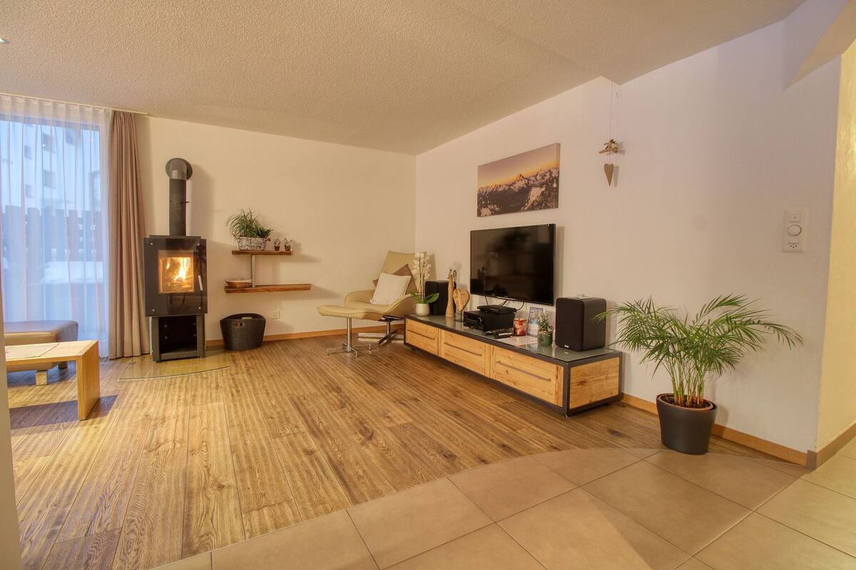 Een moderne woonkamer met een houtkachel, een flatscreen televisie, een bank, een houten zitbank en een potplant.