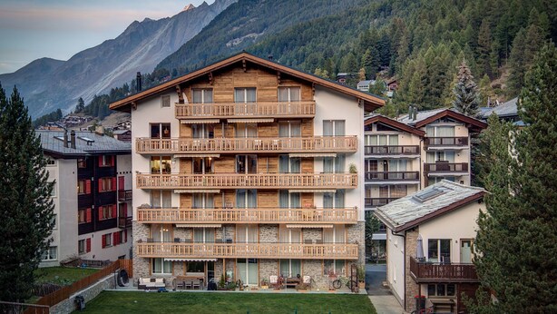A multi-story hotel with wooden balconies, a stone facade, and a view of mountains. - Matterhorngruss Apartments (Zermatt)
