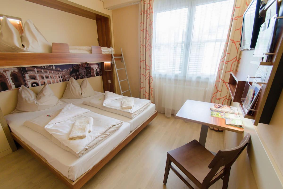 A double bed with pillows and a blanket, a wooden desk with a chair, and a window with curtains.