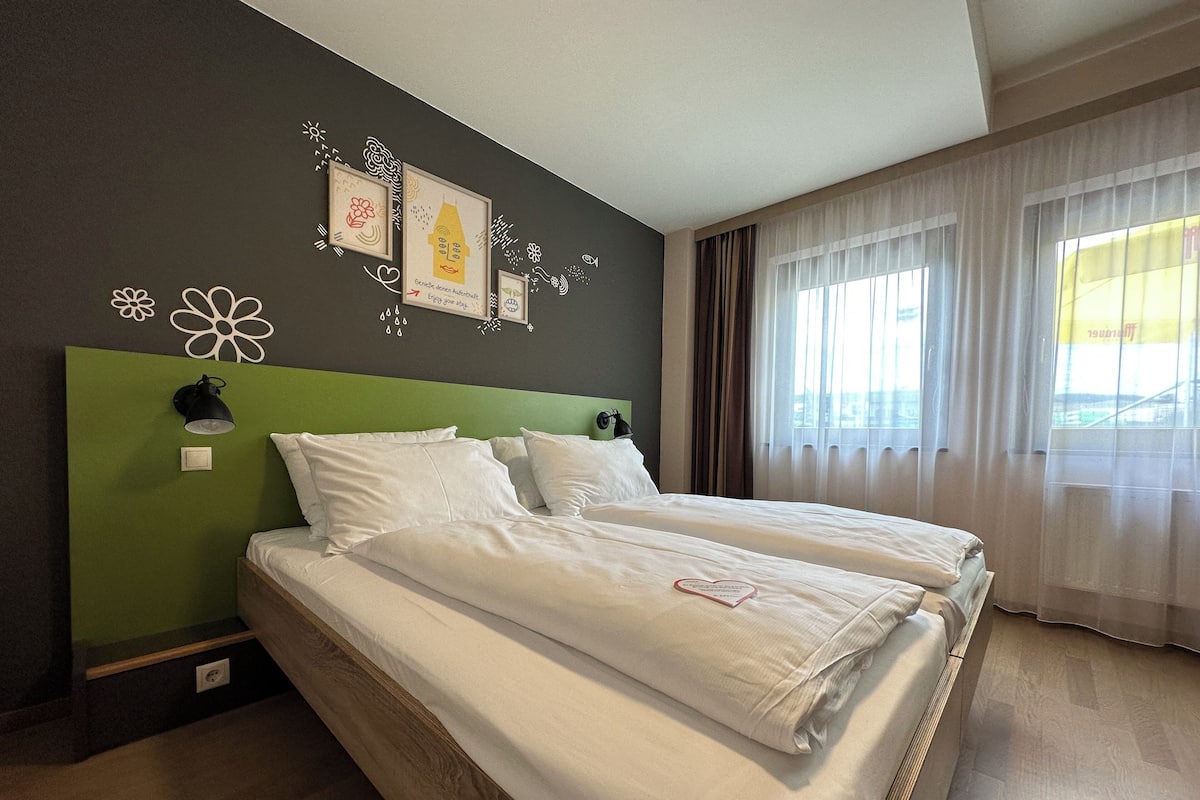 A double bed with white linens in a room with a green accent wall, a wall-mounted lamp, and a window with sheer curtains.
