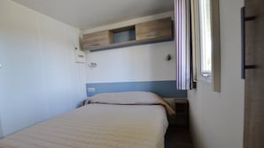 Una cama individual con colcha color beige, una mesita de noche pequeña, una ventana con cortinas y un estante empotrado sobre la cama.