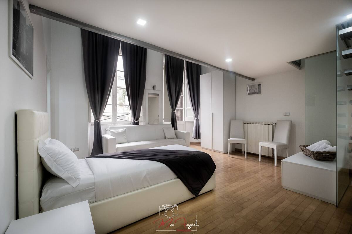 A modern hotel room with a large bed, a sitting area with a sofa and chairs, a glass partition, and a small table with a basket.