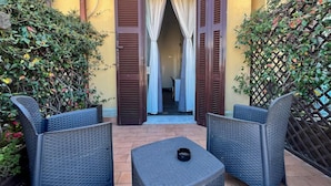 An outdoor seating area with wicker furniture and a small table, surrounded by plants and a yellow building with open shutters. - Arch Rome Suites (Rome)