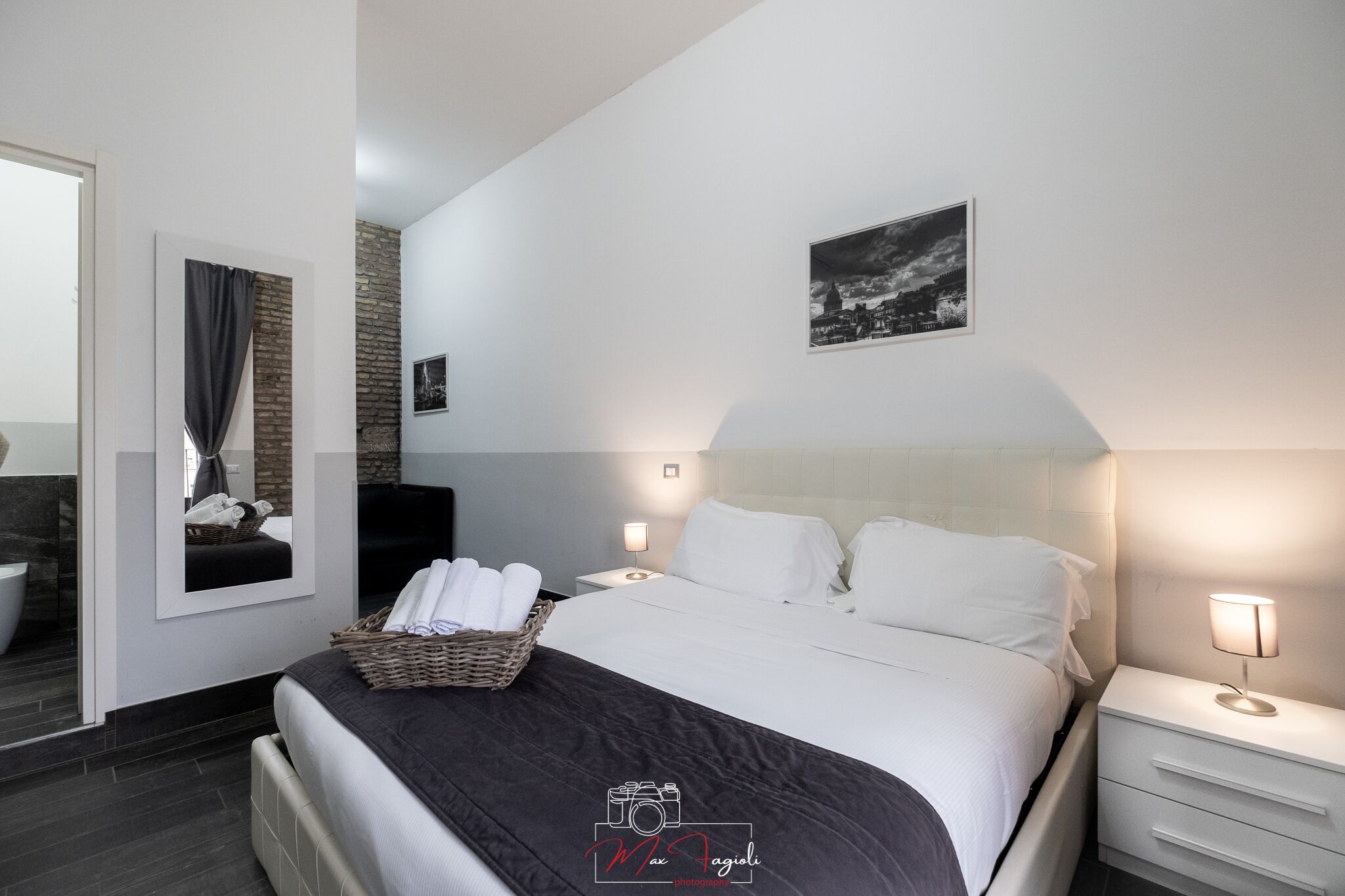A modern hotel room with a large bed, a nightstand, a wall-mounted lamp, a framed picture, and a basket of towels.