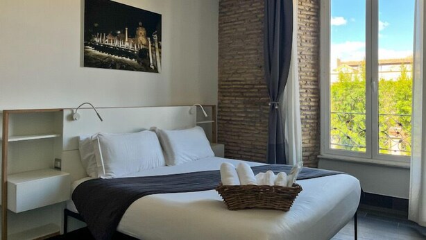 A hotel room with a large bed, a headboard, a nightstand, a window with a view of greenery, and a wall-mounted picture. - Arch Rome Suites (Rome)