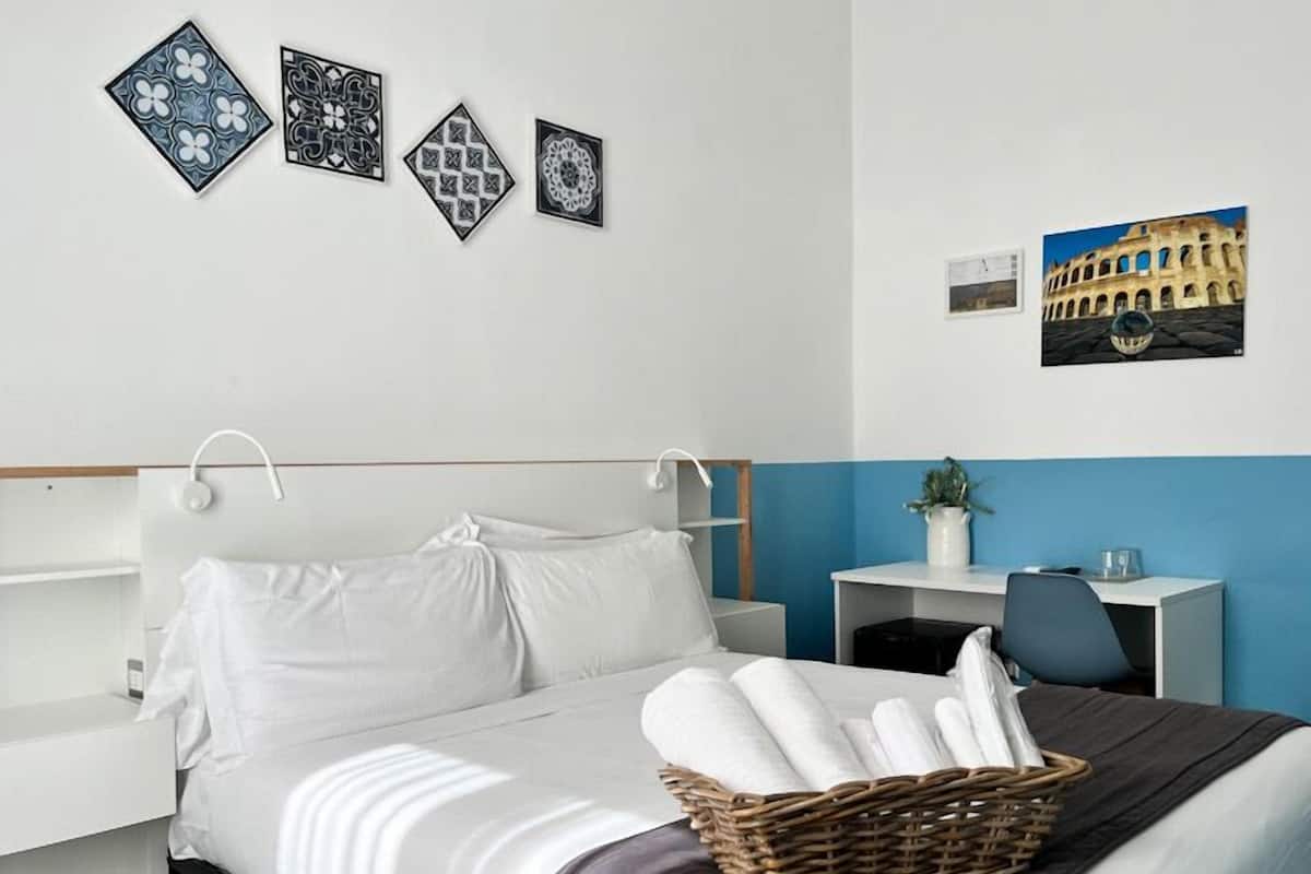 A hotel room with a bed, a desk, a chair, and a wall with decorative tiles and a picture of the Colosseum.