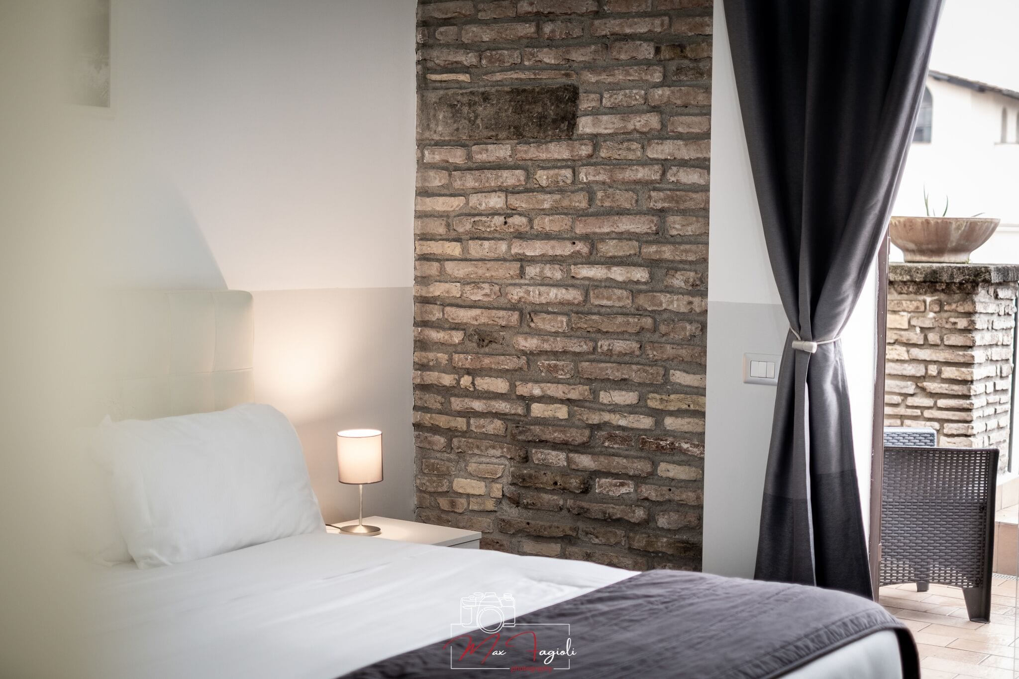 A bedroom with a brick wall, a bed with white bedding, a nightstand with a lamp, and a view of an outdoor area with a wicker chair.