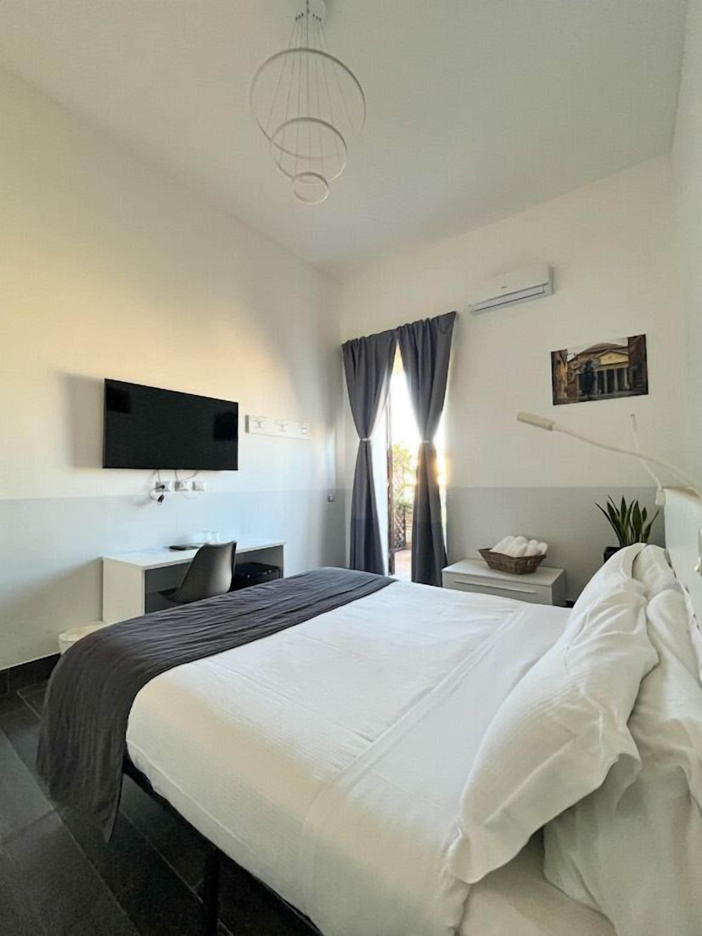 A modern hotel room with a large bed, a desk, a flat-screen TV mounted on the wall, and a window with curtains.