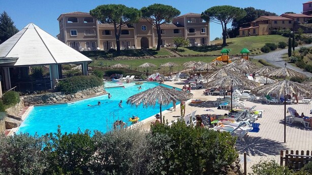 A swimming pool surrounded by lounge chairs and umbrellas, with a building complex in the background.