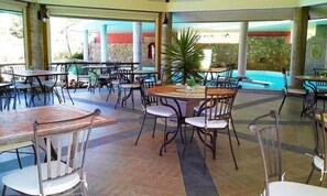 A covered outdoor dining area with multiple tables and chairs, a central decorative plant, and a view of the outside.