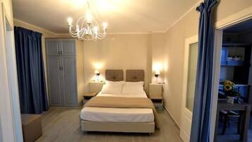 A hotel room with a large bed, two bedside tables with lamps, a chandelier, a wardrobe, and a view of the dining area.