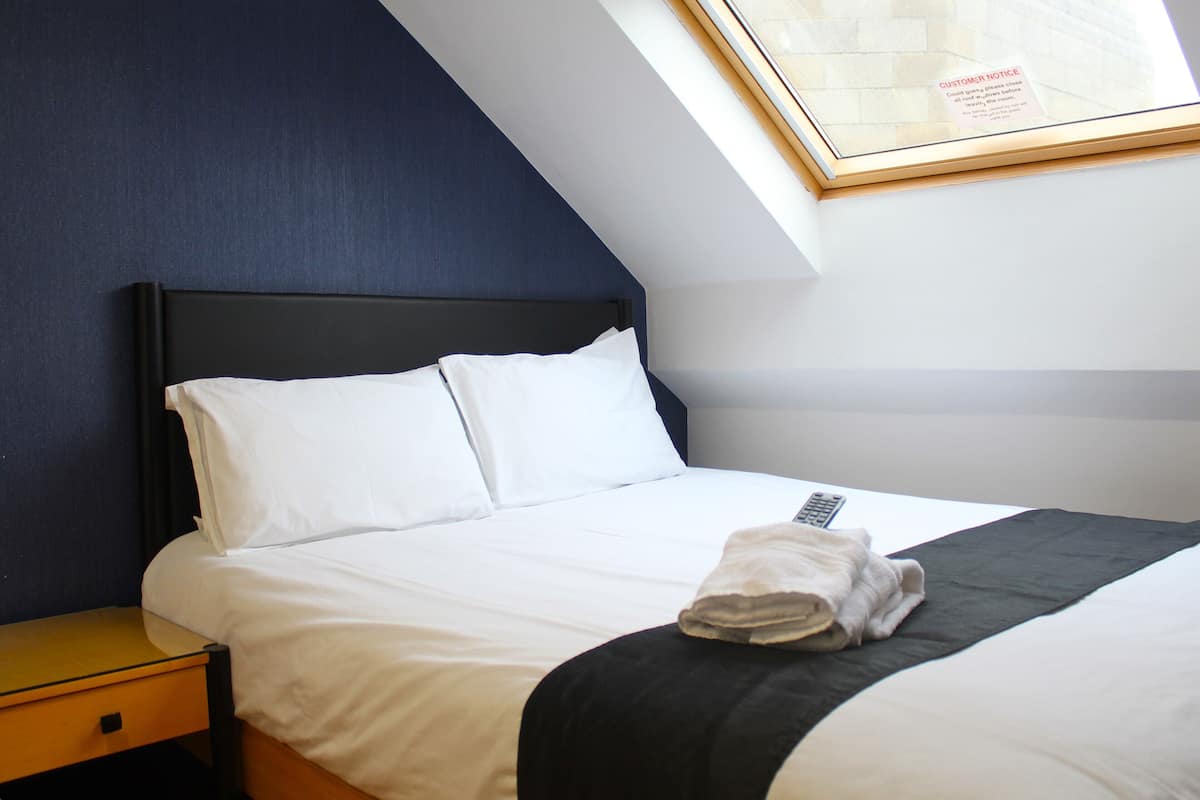 Een net opgemaakt bed met witte lakens en een zwart plaids, een nachtkastje en een ingelijst bericht aan de muur.