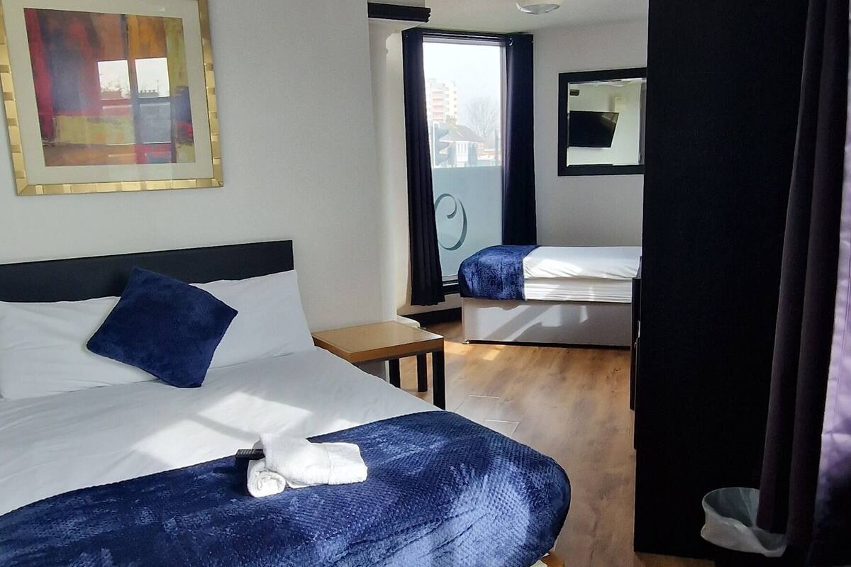 Een hotelkamer met houten vloer, een bed met blauwe deken, een televisie en een raam met uitzicht op gebouwen.