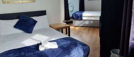 A hotel room with a wooden floor, a bed with a blue blanket, a TV, and a window with a view of buildings.