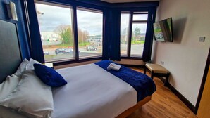 Una habitación de hotel con una cama grande, un televisor fijado en la pared y vistas a la calle a través de amplias ventanas.