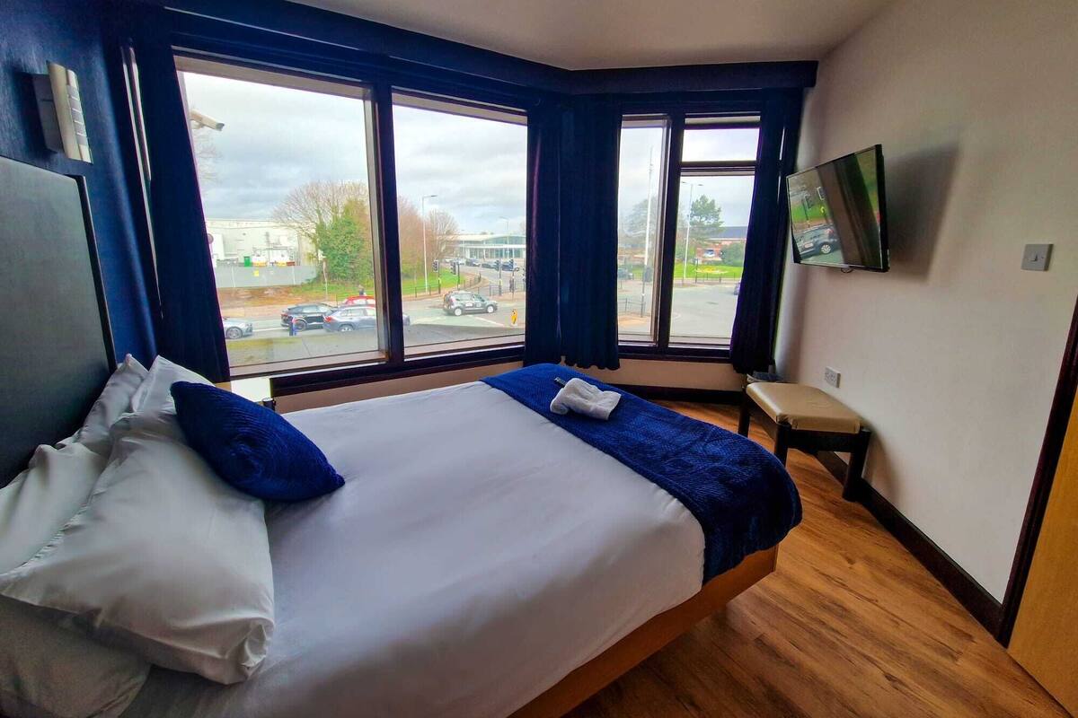 Een hotelkamer met een groot bed, een aan de muur bevestigde televisie en uitzicht op de straat door grote ramen.