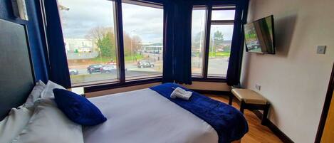 Una habitación de hotel con una cama grande, un televisor fijado en la pared y vistas a la calle a través de amplias ventanas.