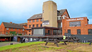 Un hotel con una torre de esquina destacada, un restaurante llamado 'Osteria' en un lateral y un área de picnic en primer plano.