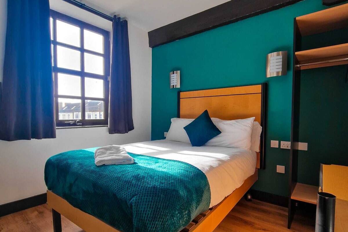 Een hotelkamer met een houten bed, wanden in blauwgroen, een raam met blauwe gordijnen en uitzicht op gebouwen.