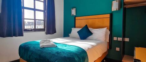 Quarto de hotel com cama de madeira, paredes verde-água, janela com cortinas azuis e vista para edifícios.