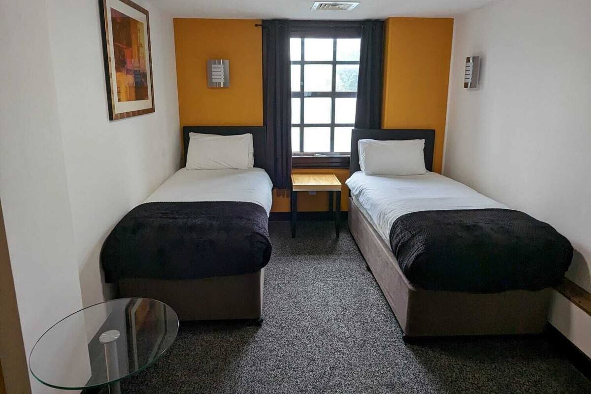 Een hotelkamer met twee eenpersoonsbedden, een tafeltje, een raam met gordijnen en een plafondlamp.
