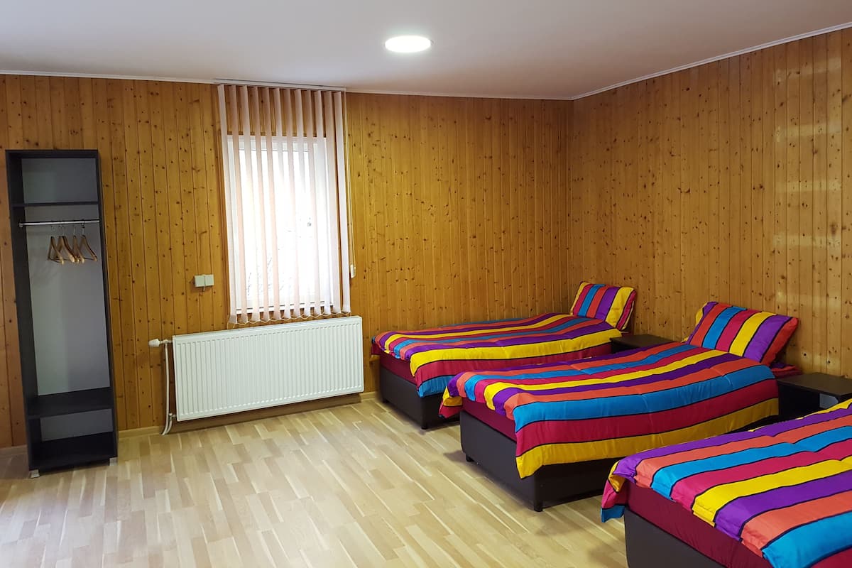 Quarto com paredes e soalho de madeira, duas camas com roupa de cama colorida listrada, um radiador, um guarda-roupa e uma janela com persianas.