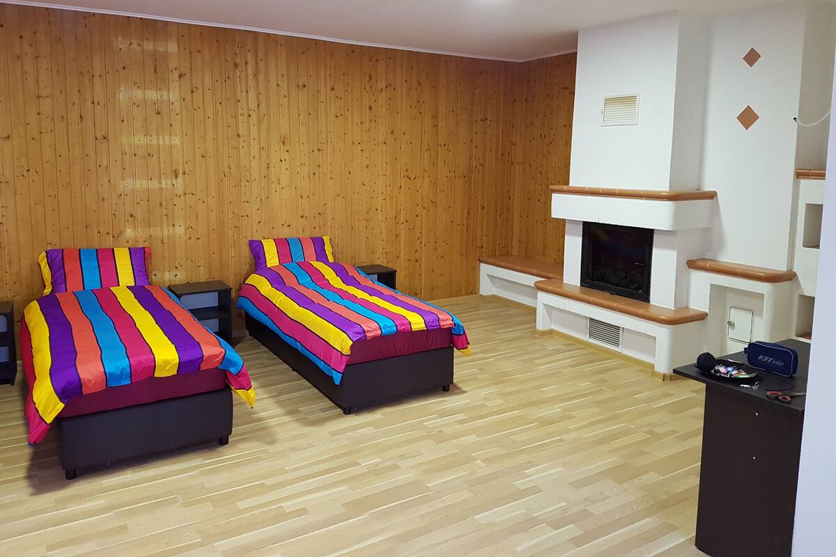 Quarto com duas camas, parede de madeira, lareira e uma pequena escrivaninha.