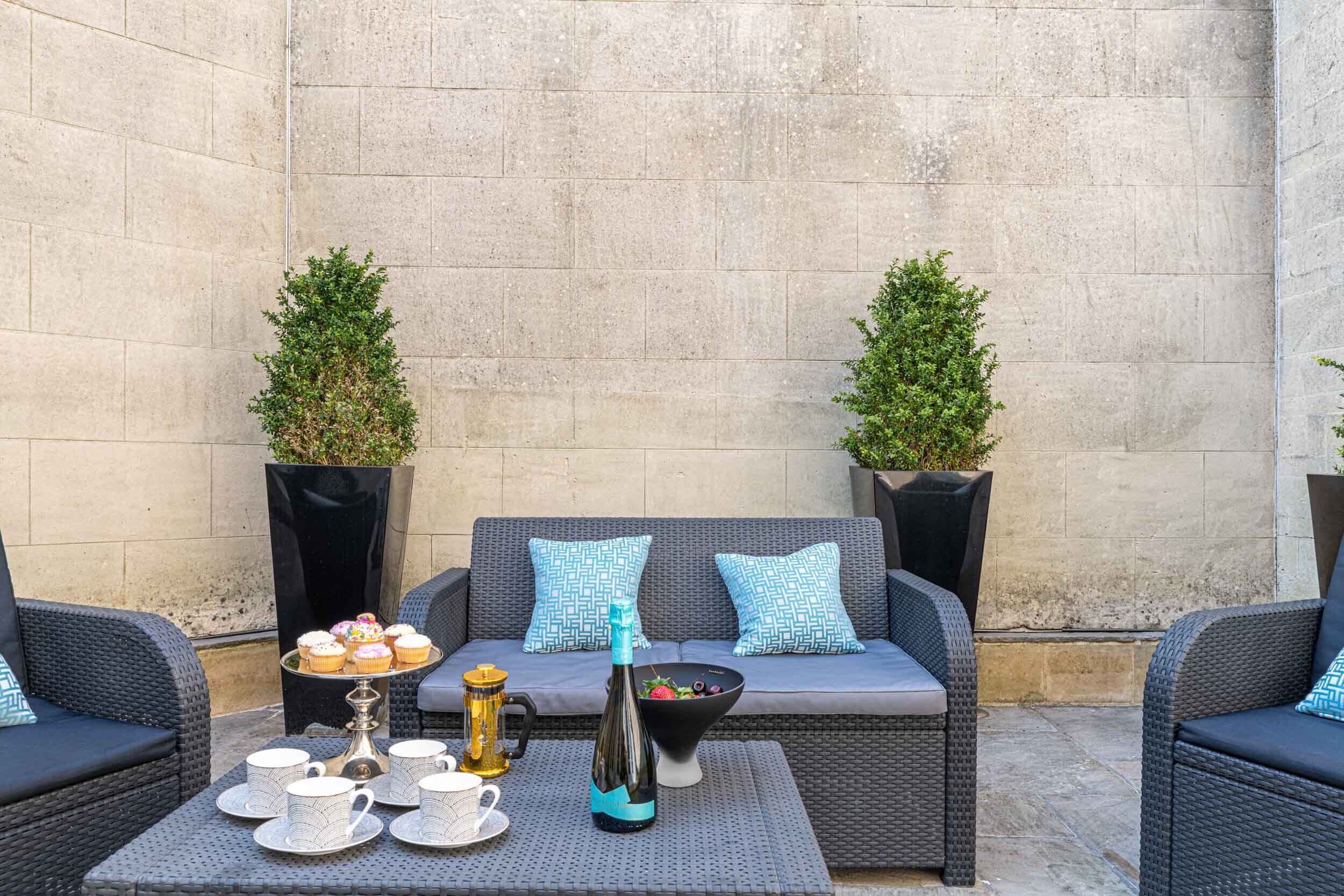An outdoor seating area with a grey sofa, two potted plants, a table with a bottle, a tray of cupcakes, and a bowl of fruit.