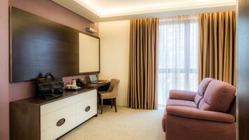 A hotel room with a large mirror, a desk with a laptop, a chair, a pink sofa, and a large window with curtains.