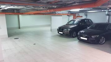A parking garage with two cars parked, white walls, and visible pipes and ducts on the ceiling.