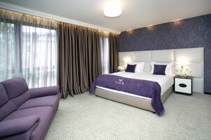A hotel room with a large bed, a sofa, a nightstand with a lamp, and a view of the outdoors through the window.