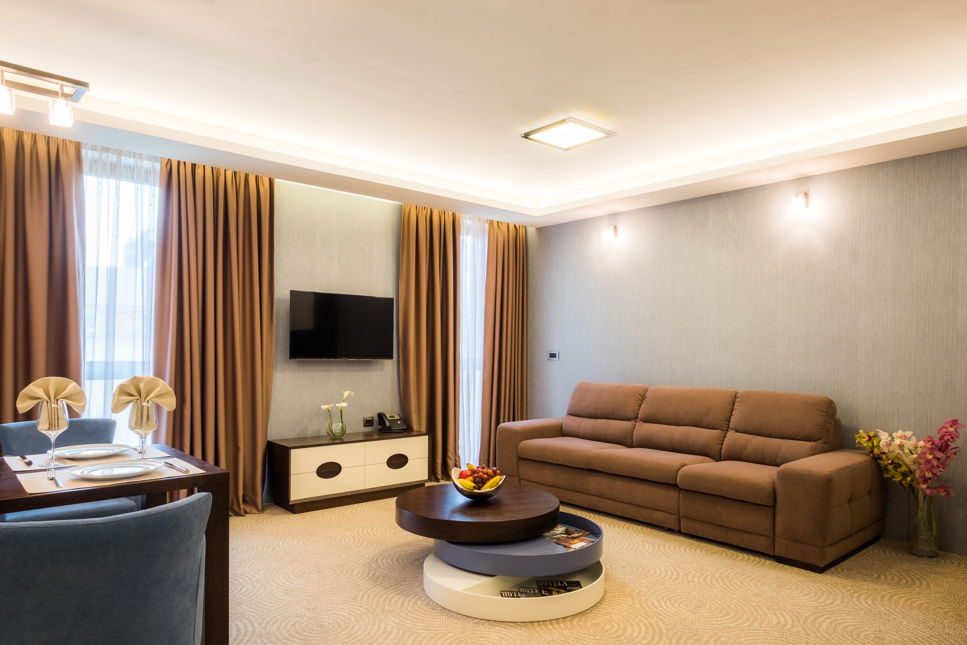 Ein modernes Hotelzimmer mit einem braunen Sofa, einem Esstisch für zwei Personen, einem wandmontierten Flachbildfernseher und einer dekorativen Vase mit Blumen.