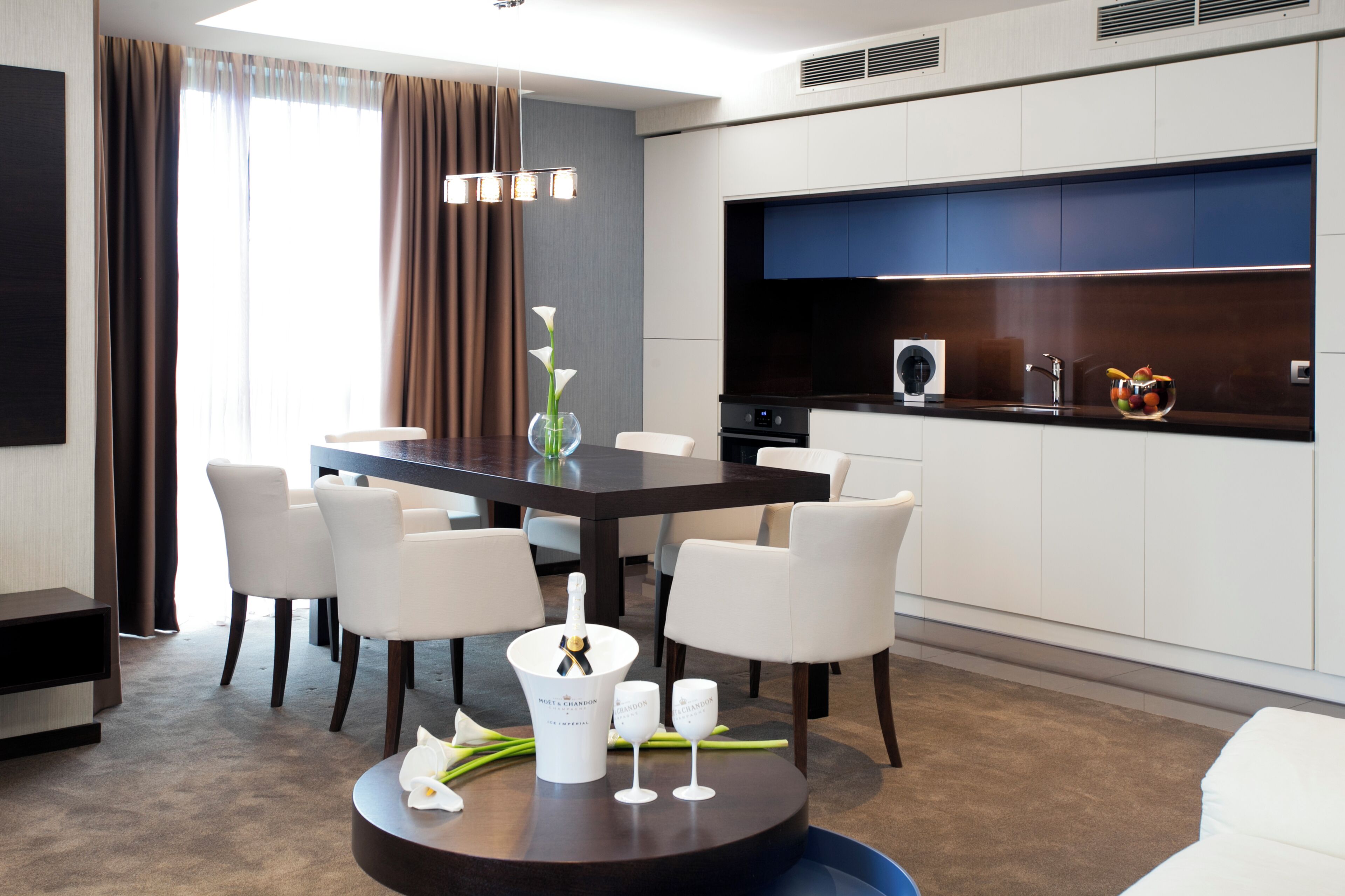 A modern dining area with a dark wooden table, white chairs, a vase with flowers, and a bottle of champagne.