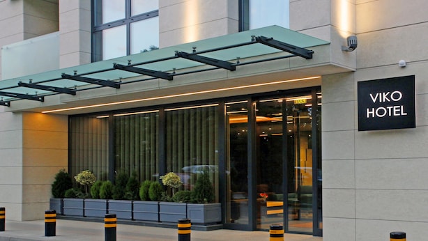 Una entrada de hotel moderna con fachada de vidrio, un cartel que dice "VIKO HOTEL" y un área exterior cubierta con plantas.